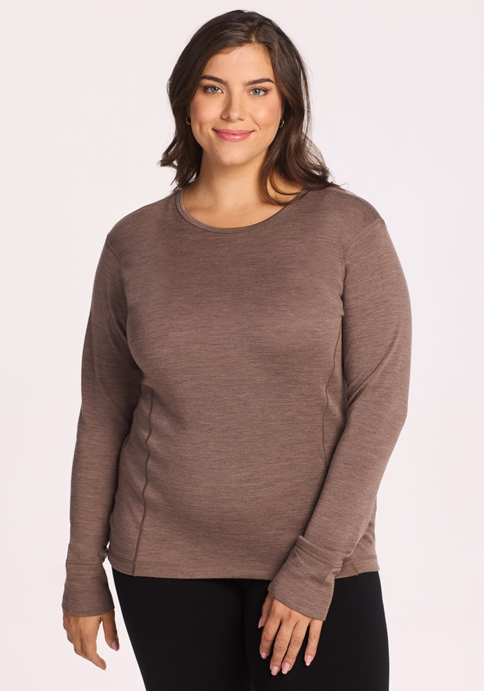 A woman with long brown hair, wearing the Woolx Hannah Long Sleeve Top in Simply Taupe and black pants, stands facing the camera and smiles gently against a plain light background. 
