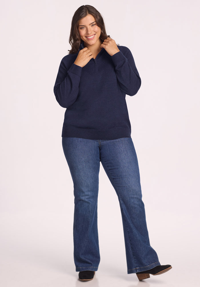 A woman with long brown hair smiles while wearing the Woolx Collins Half Zip Sweater in Storm Cloud, paired with blue flared jeans and black boots, against a plain white background.