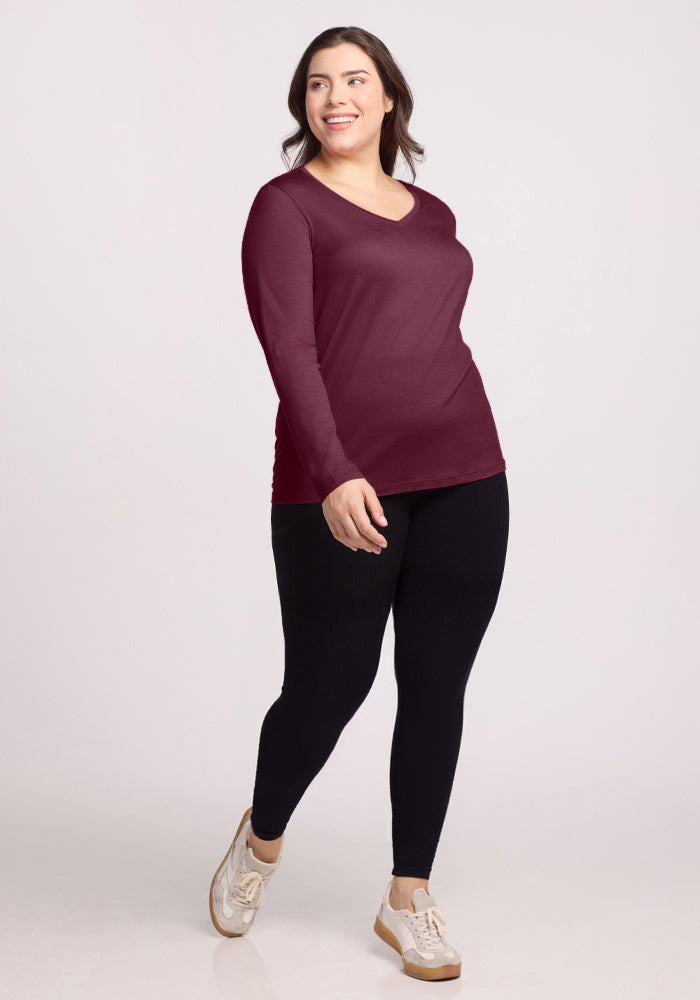 A woman smiles and walks against a plain light background, wearing the Woolx Millie Long Sleeve in Grape Harvest, black leggings, and white sneakers.
