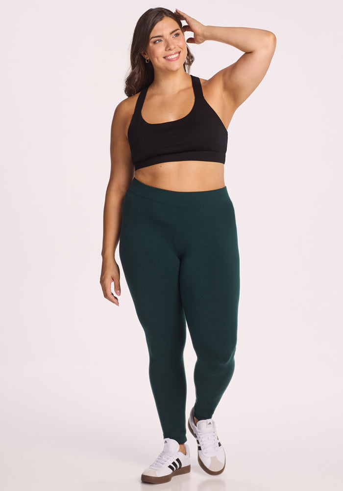 A woman with long brown hair wears the Woolx Stella Tall - Deep Timber heavyweight leggings in dark green, a black sports bra, and white sneakers with black stripes. She smiles and touches her hair while standing against a light background.