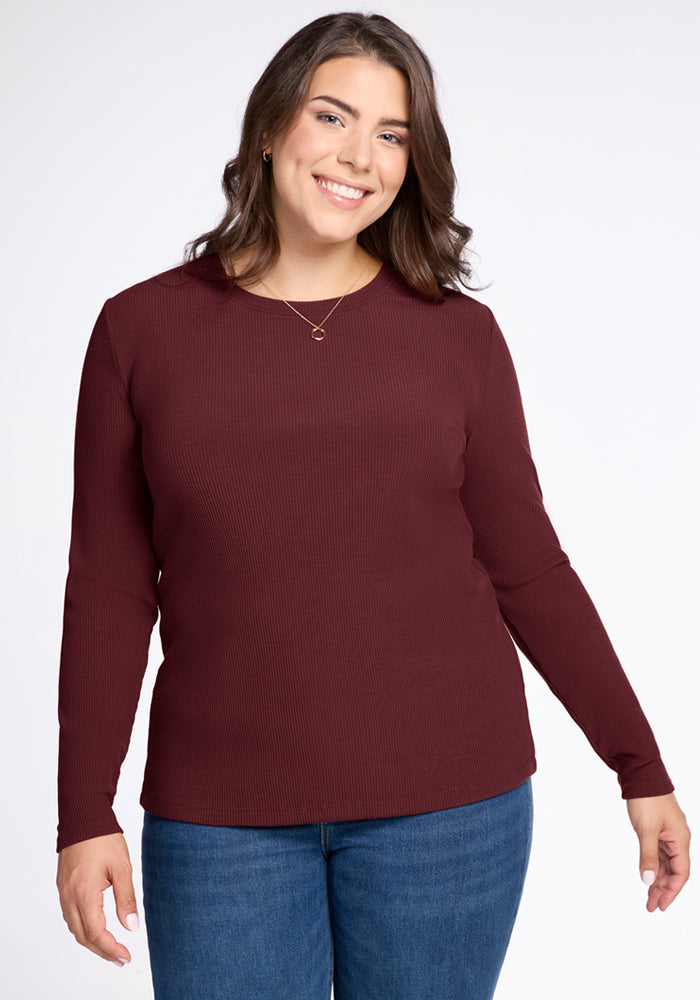 A woman with long brown hair wears the Woolx Hadley Ribbed Crew in Toasted Garnet and blue jeans, smiling at the camera against a plain light background—perfect for lightweight layering and travel. 
