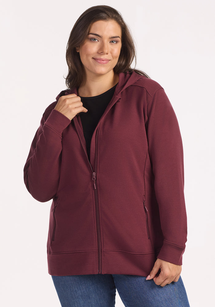 A woman with long brown hair smiles while wearing the Woolx Darby Zip Up Hoodie in Toasted Garnet over a black shirt and blue jeans, standing against a plain light background. 