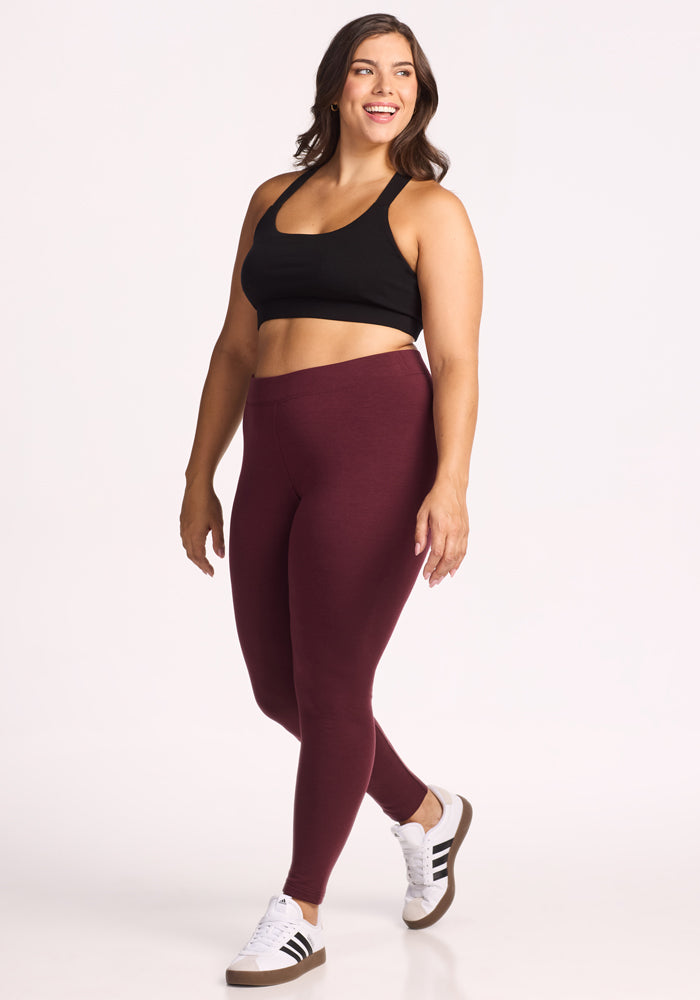 A woman smiles while walking in a black base layer sports bra, Woolx Stella Leggings in Toasted Garnet, and white sneakers with black stripes, set against a plain white background.