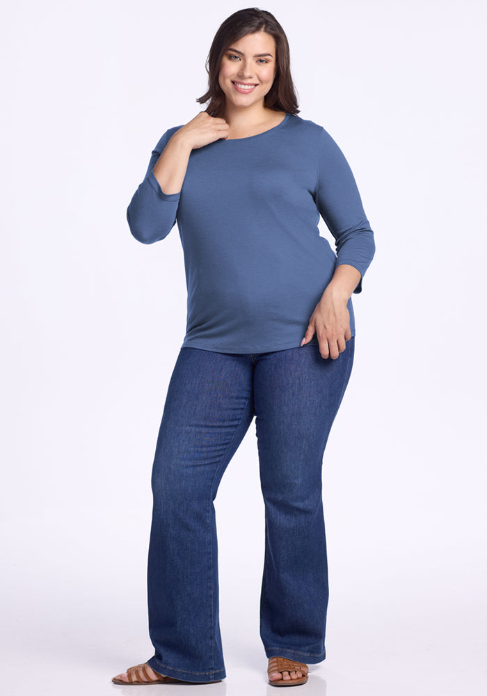 A smiling woman with dark hair wears the Woolx Jenny 3/4 Sleeve Crew Neck in Skyline, paired with dark blue jeans and brown sandals, standing against a plain white background with one hand near her neck and the other on her hip.