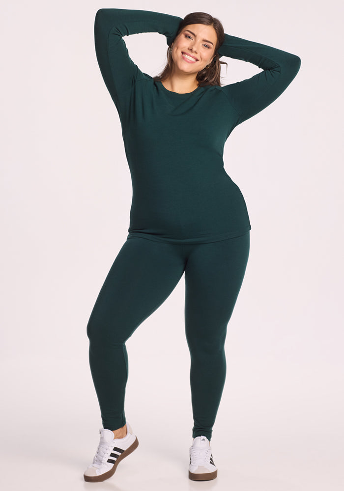 A smiling woman poses with her hands behind her head, wearing the Woolx Sadie Baselayer Top in Deep Timber and matching leggings with white sneakers, standing against a plain light background.