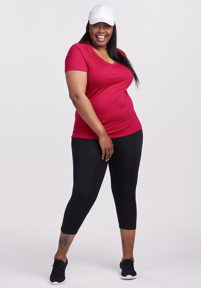 A smiling woman wears the Woolx Mia V Neck in Poppy Spark, a white cap, black leggings, and black sneakers as she poses confidently against a plain white background.