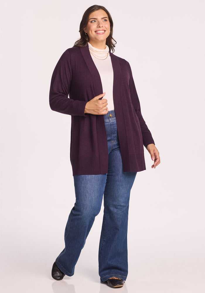 A woman smiles wearing the Woolx Macie Sweater in Deep Plum over a white turtleneck, paired with blue flared jeans and black shoes, standing against a plain light background.