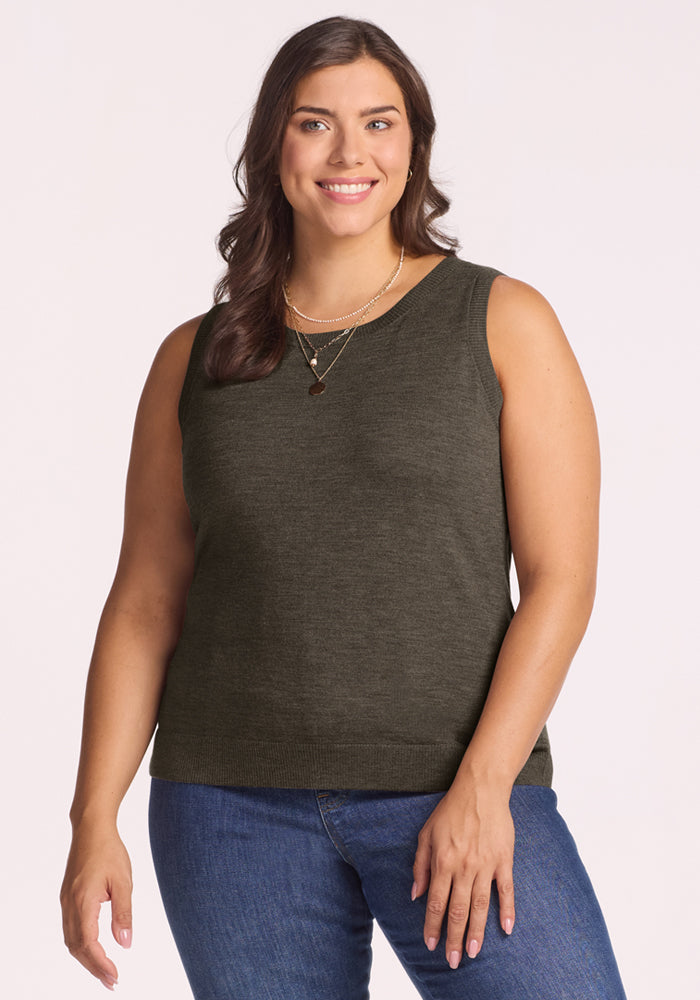A woman with long brown hair wears the Woolx Chessy Tank Top in Pine Needle, blue jeans, and layered necklaces, smiling against a plain light background. 