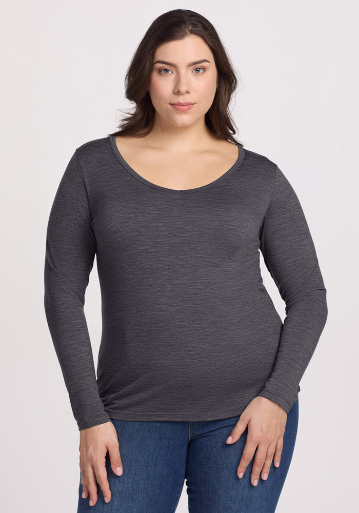 A woman with long brown hair wears the Woolx Layla V Neck Top in Cinder Melange and blue jeans, standing against a plain light background, facing the camera. 