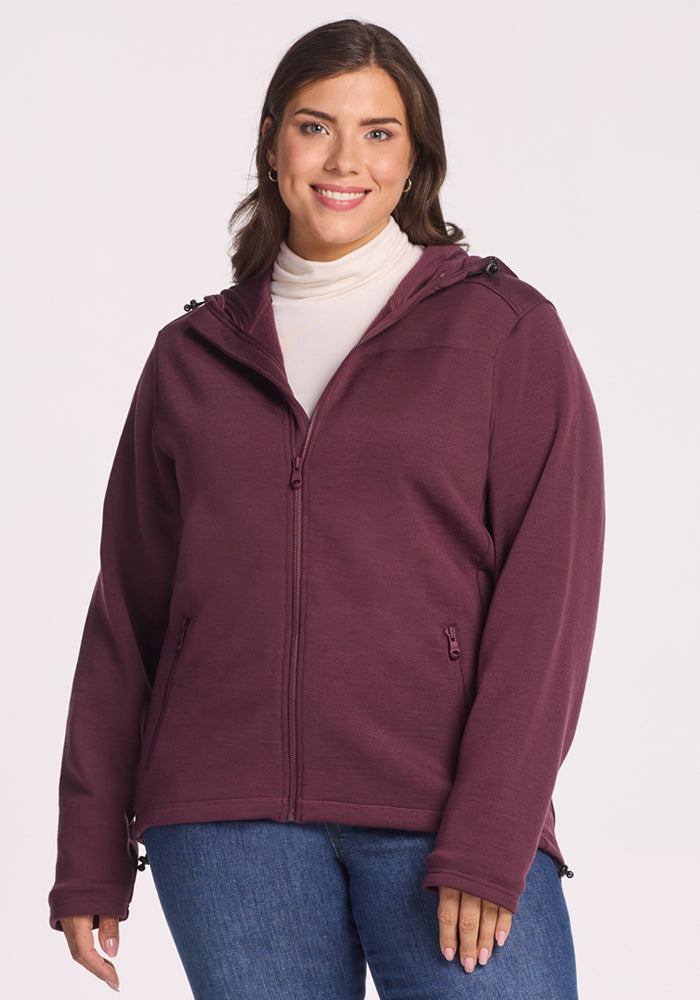 A woman smiles against a plain light background wearing the Woolx Cubby Hooded Sweatshirt in Grape Harvest over a white turtleneck and blue jeans. 