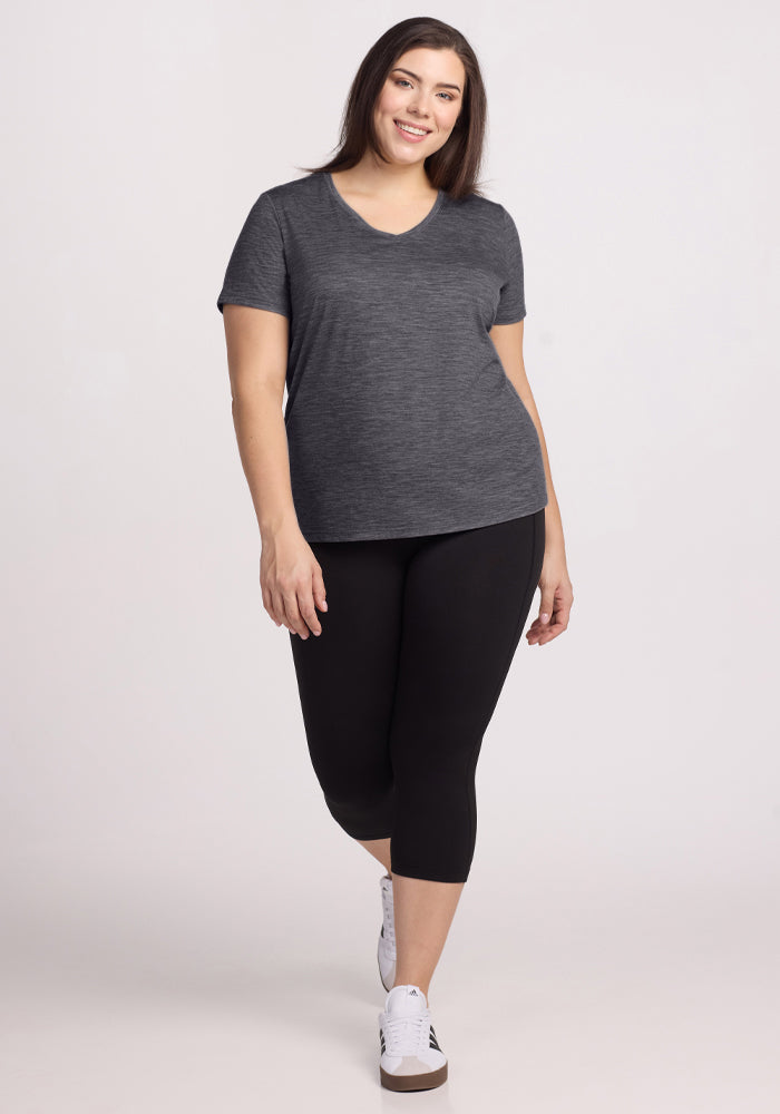 A woman with long dark hair smiles while wearing the Woolx Junie V Neck - Cinder Melange, paired with black capri leggings and white sneakers. The moisture-wicking Merino wool keeps her comfortable and stylish against a light background.