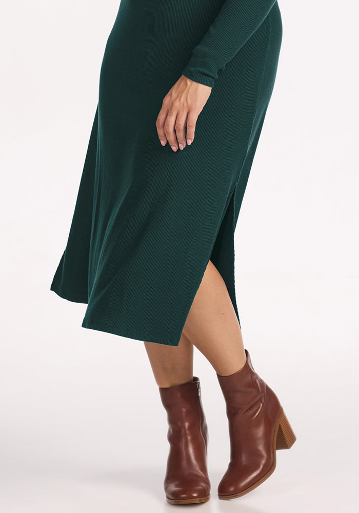 A person wearing the Woolx Caroline Ribbed Twirl Dress in Deep Timber with brown heeled ankle boots, standing against a plain white background.