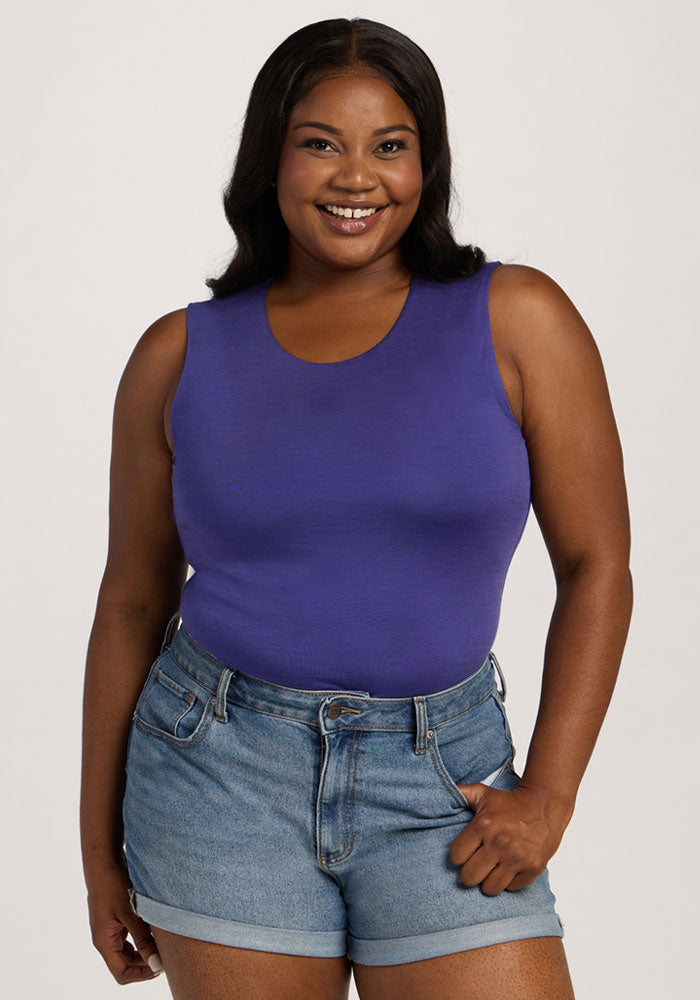 Smiling woman in the Woolx Maggie Bodysuit - Berry Indigo and denim shorts, standing against a plain light background. 