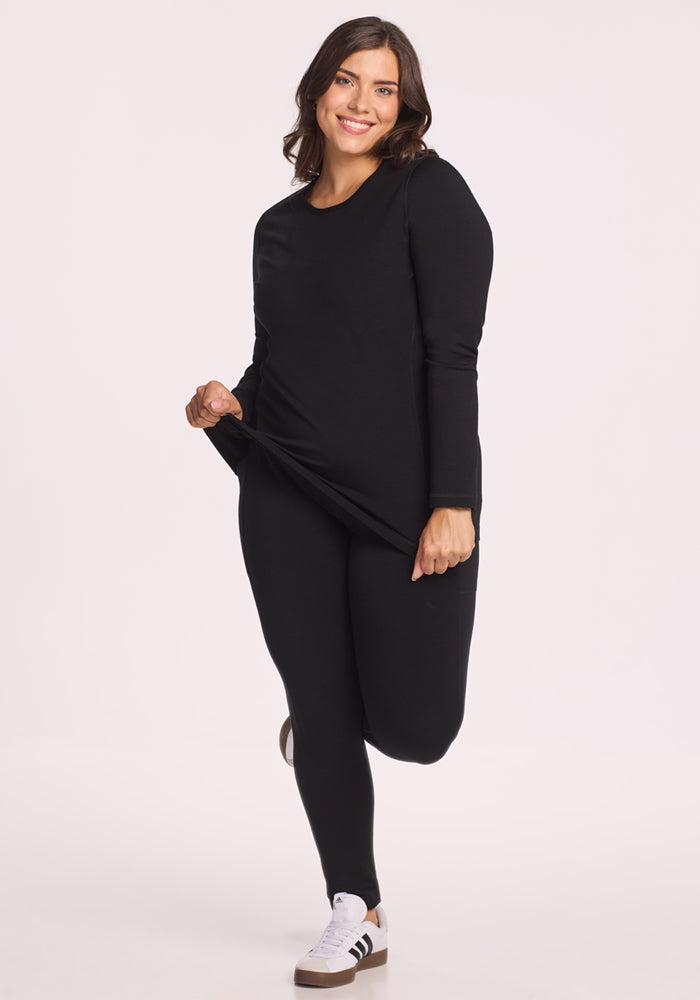 A woman wearing the Woolx Elsa Long Sleeve - Black and matching leggings stands on one leg, lifting one side of her top and smiling at the camera. She pairs the outfit with white sneakers featuring black stripes.