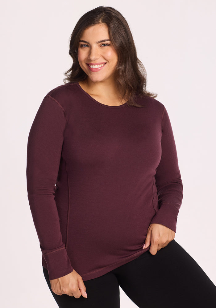 A woman with long, dark hair smiles while wearing the Woolx Hannah Long Sleeve Top in Chocolate Fig and black pants against a plain, light background. 