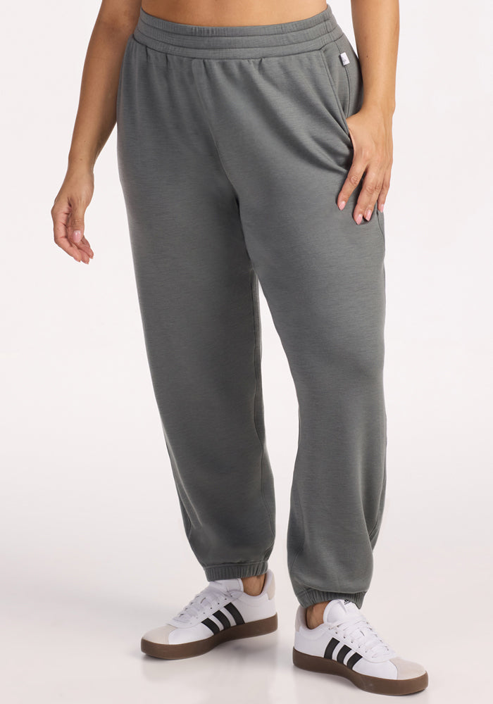 A person wears Woolx Parker Sweatpants in Sedona Sage, paired with white sneakers with black stripes, standing against a plain light background. The moisture-wicking fabric keeps them comfortable as their left hand rests on their hip. 
