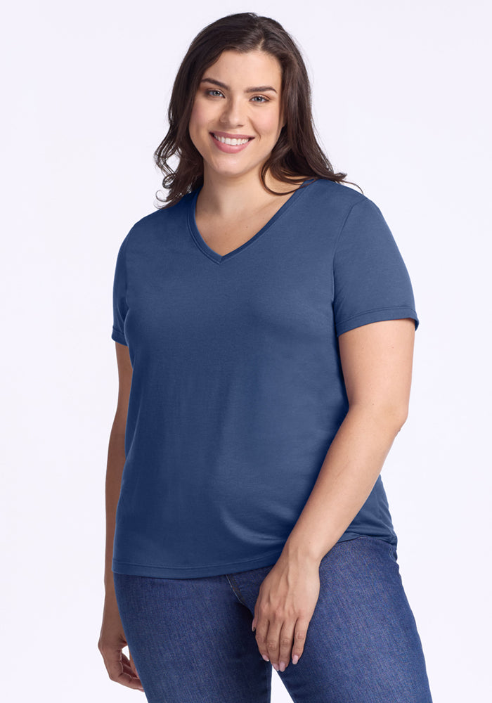 A woman with long brown hair smiles in the Woolx Junie V Neck - Starry Night, a relaxed-fit blue T-shirt, paired with blue jeans and standing against a plain light background. 