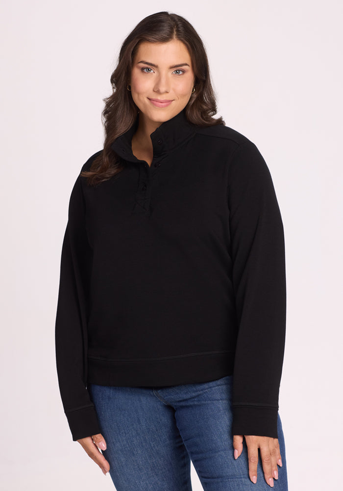 A woman with long brown hair, wearing the Woolx Annie 3 Button Pullover in black and blue jeans, stands against a plain light background and smiles softly with one hand by her side. 