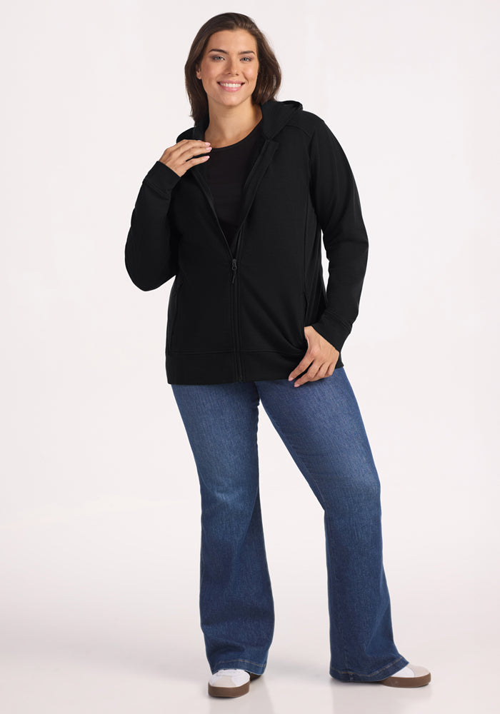 A woman with brown hair smiles while wearing the Woolx Darby Zip Up Hoodie in black, paired with a black top and blue jeans. She stands against a plain white background, with one hand in her pocket and the other holding the hoodie.