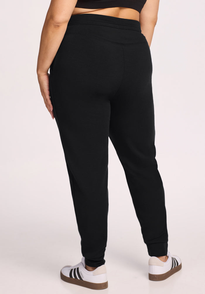 A person wearing Woolx Willa Joggers in black, a black crop top, and white sneakers with black stripes stands on a light-colored background, shown from the back.