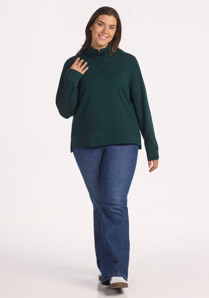 A woman with long brown hair smiles as she walks forward, wearing the Woolx Annie 3 Button Pullover in Deep Timber, paired with blue jeans and white shoes against a plain white background.