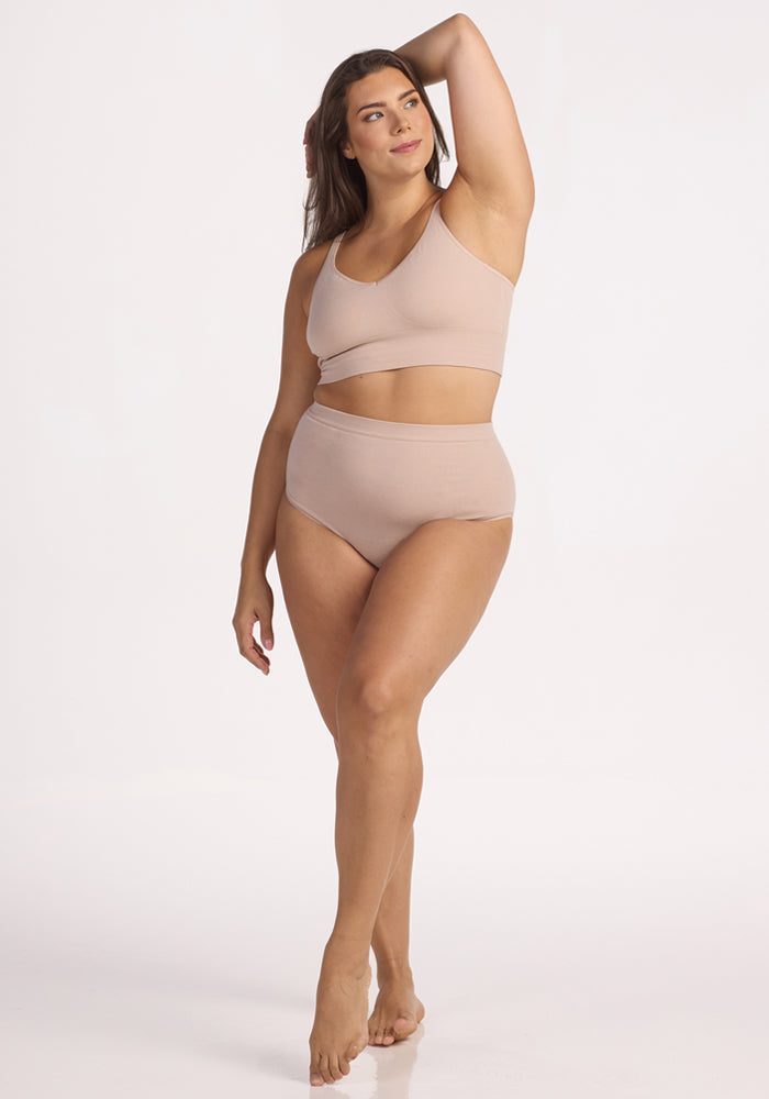 A woman poses confidently in Woolx Mabel Full Coverage Underwear – Sand, paired with a matching bralette, against a plain white background, her arm raised and expression relaxed.