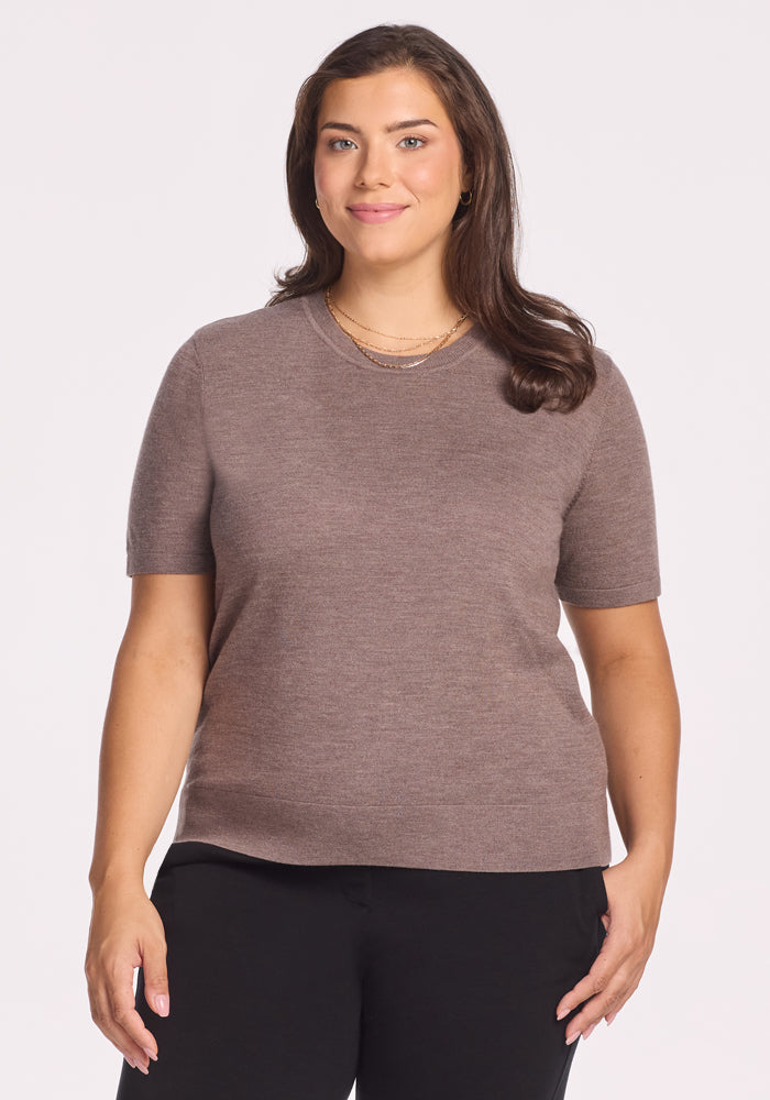 A woman with long brown hair wears the Woolx Anna Crew Sweater in Simply Taupe and black pants, smiling gently against a plain white background. 