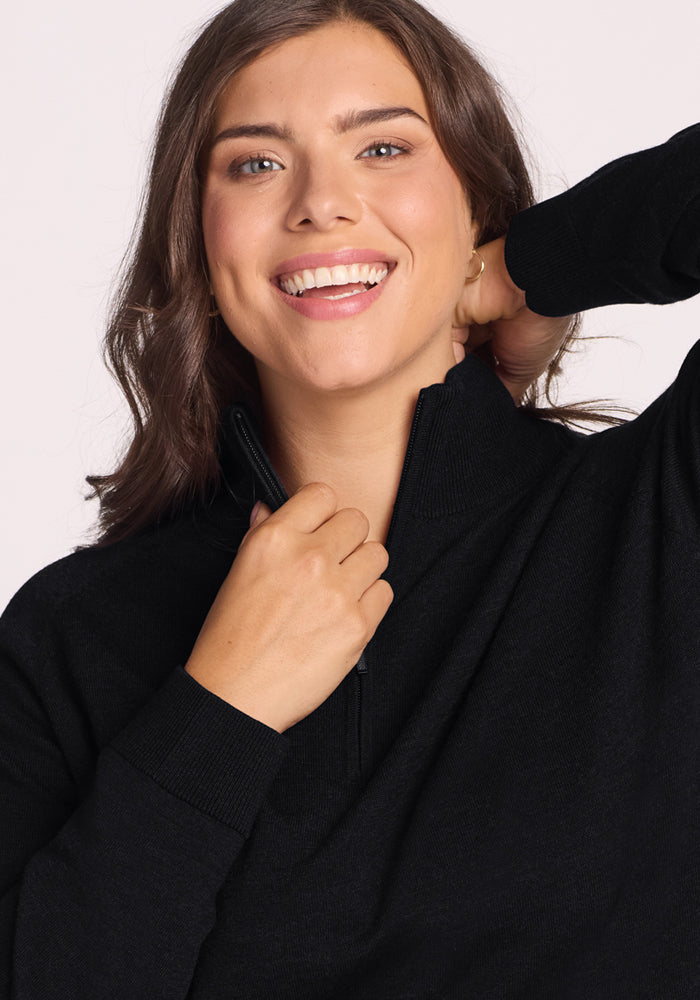A woman with wavy brown hair smiles at the camera while zipping up the Woolx Collins Half Zip Sweater in black, her arm raised to touch her neck. She stands against a light, plain background.