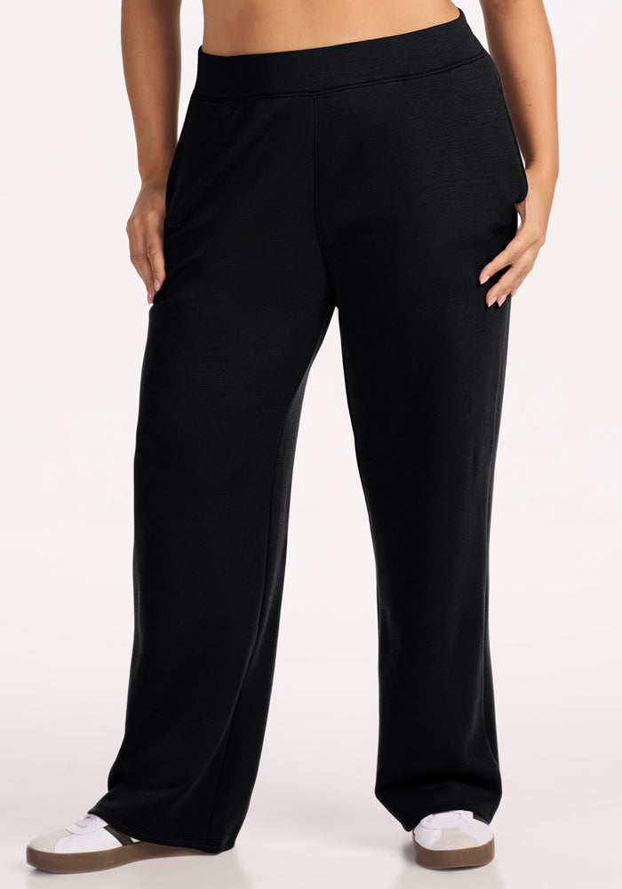 A person wears Woolx Bree Lounge Pants in black with white sneakers, standing from waist to feet with one hand in a pocket against a plain light background.