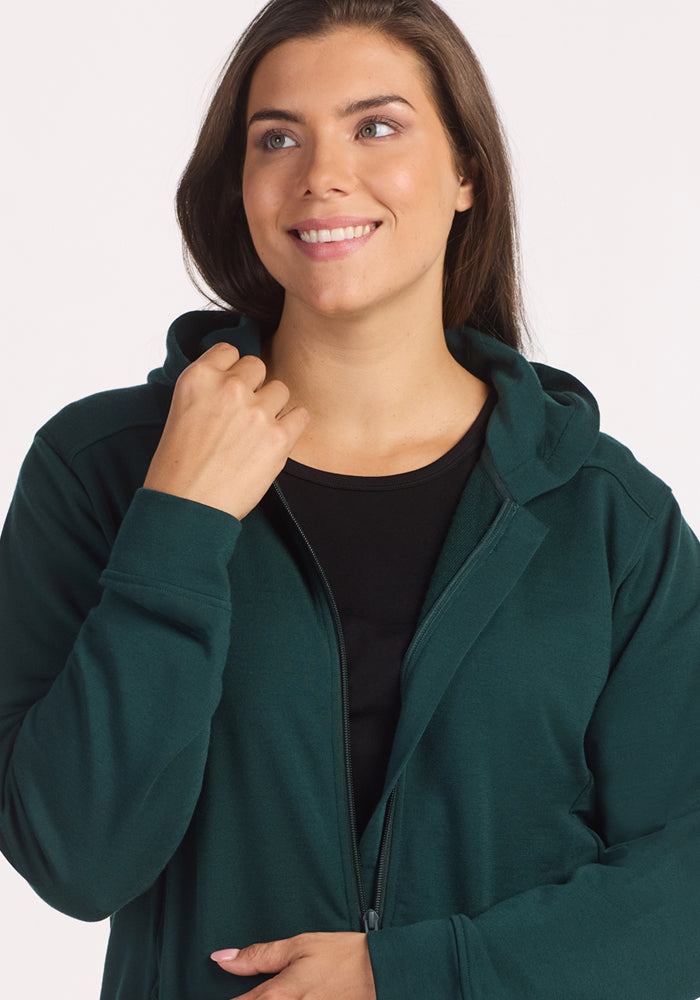 A woman with long brown hair smiles and looks upward while wearing the Woolx Darby Zip Up Hoodie in Deep Timber over a black top, holding the hoodie near her neck. The background is plain white.