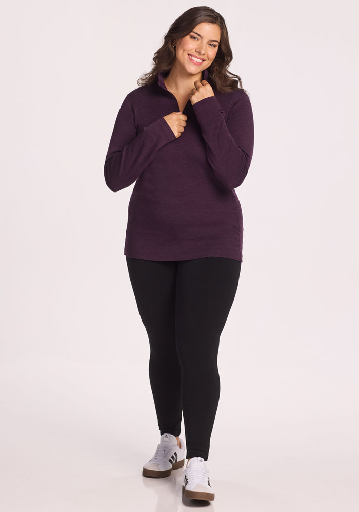 A smiling woman with long dark hair wears the Woolx Brooke 1/4 Zip in Deep Plum, paired with black leggings and white sneakers, standing against a plain light background.