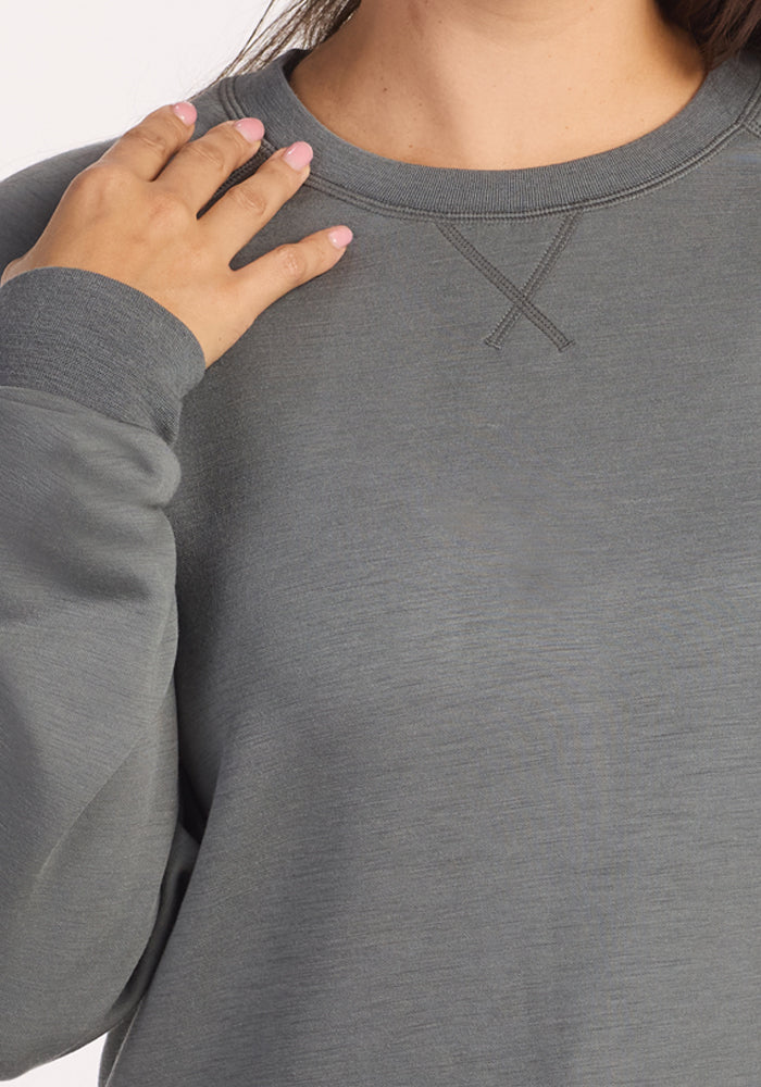 A person wearing the Woolx Bailey Crewneck Sweatshirt in Sedona Sage touches their left shoulder with their right hand. Only their lower face, neck, and upper torso are visible, and their nails are painted light pink.