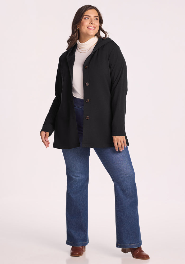 A woman with long brown hair wears the Woolx Edna Hooded Cardigan in black over a white turtleneck, blue jeans, and brown boots. She is smiling and standing against a plain light background.