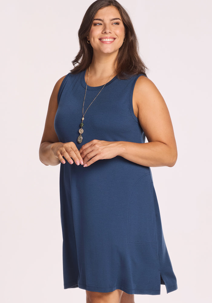A woman with long brown hair smiles while wearing the Woolx Clara Dress - Starry Night, a sleeveless, knee-length blue summer dress. She stands against a plain light background, accessorized with a long pendant necklace.