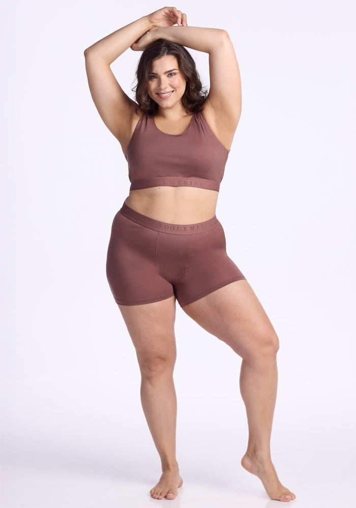 A curvy woman stands barefoot against a white background, smiling with arms raised while wearing the Woolx Elliott Boyshort in Nutmeg and a matching sports bra.