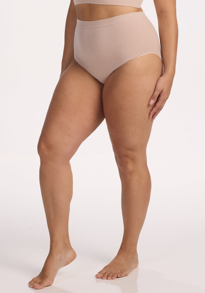 Wearing Woolx Mabel Full Coverage Underwear in Sand, a person stands barefoot against a white background, with one arm relaxed by their side. The cropped photo emphasizes the seamless comfort and modern silhouette of the design.
