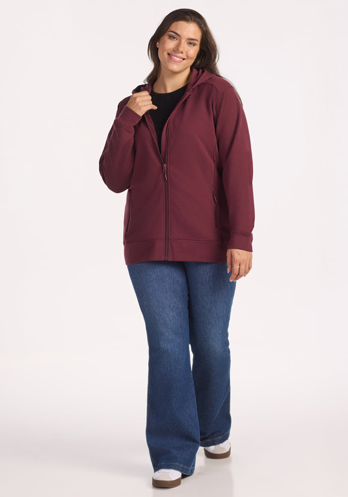 A woman smiles while wearing the Woolx Darby Zip Up Hoodie in Toasted Garnet, paired with blue jeans and white shoes. She stands against a plain white background, holding the hoodie collar with one hand.