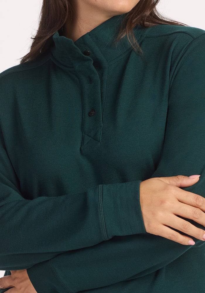 A person with long brown hair wears the Woolx Annie 3 Button Pullover in Deep Timber, arms crossed. The image is cropped from shoulders to mid-torso and set against a plain, light background.