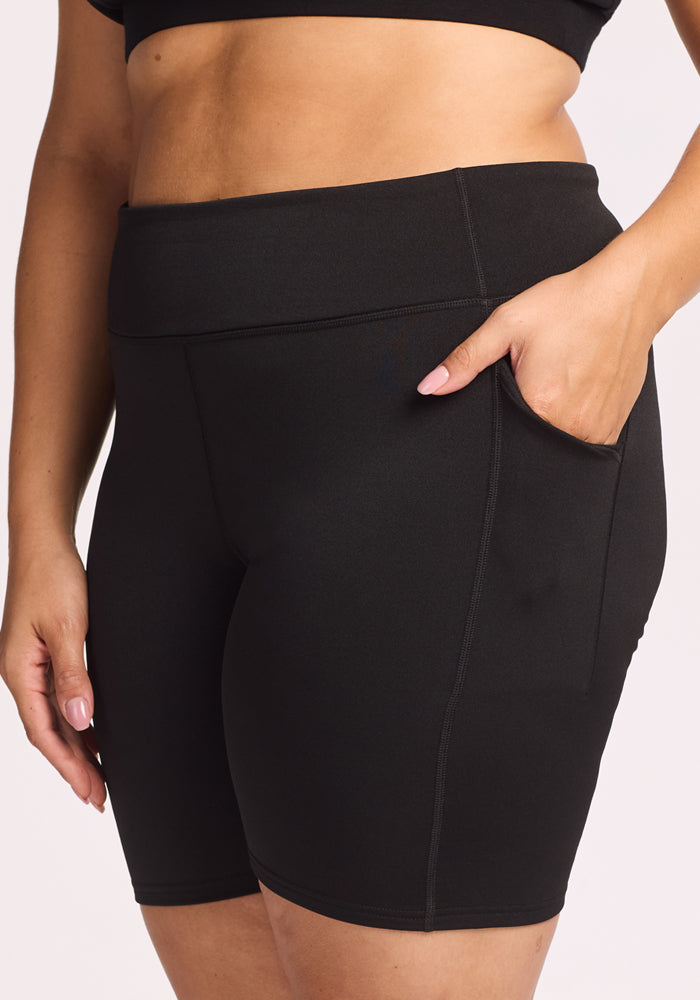 Wearing Woolx Merino Flex® Dani Bike Shorts in black, a person stands against a plain light background with one hand in the side pocket, showing off the high-waisted, moisture-wicking design.