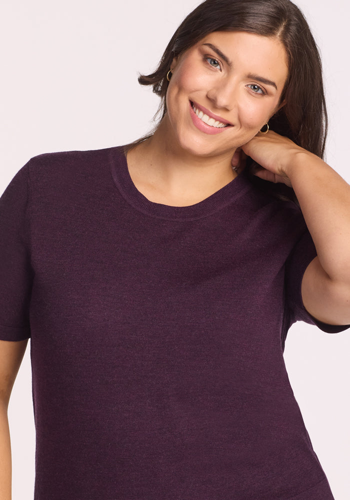 A woman with long dark hair smiles at the camera, touching her hair, while wearing the Woolx Anna Crew Sweater in Deep Plum against a plain background.