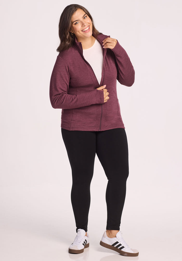 A woman smiles against a plain background, wearing the Woolx Finley Zip Up Sweatshirt in Grape Harvest Melange, paired with black leggings, a white shirt, and white sneakers with black stripes.