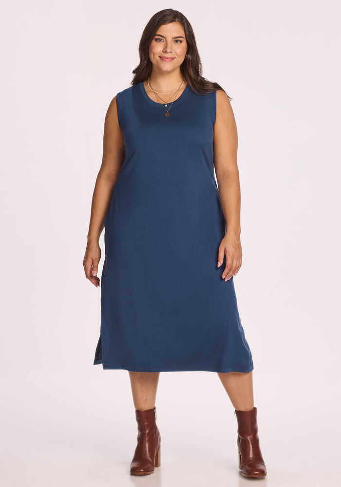 A woman with long brown hair stands smiling against a plain white background, wearing the Woolx Cassie Dress in Starry Night, a sleeveless, knee-length blue Merino wool travel dress paired with brown ankle boots. 