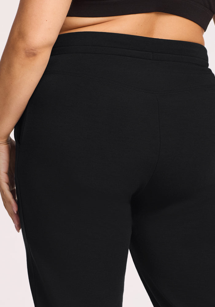 A close-up of a person wearing Woolx Willa Joggers in black and a black crop top, highlighting their midsection with one hand resting on their hip.