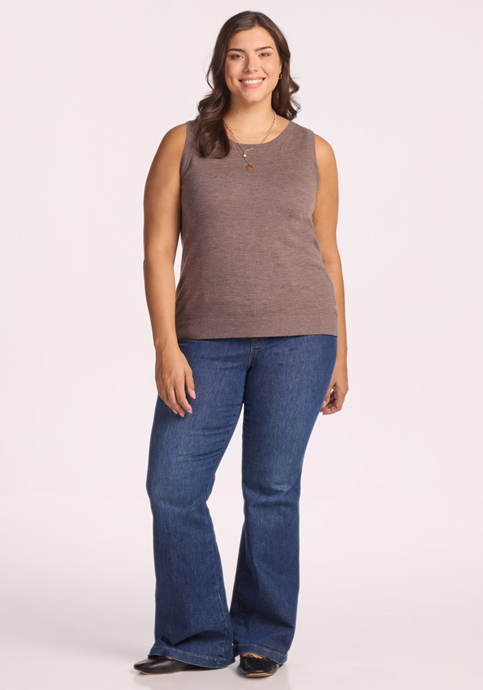 A smiling woman with long brown hair wears the Woolx Chessy Tank Top in Simply Taupe, paired with blue flared jeans and black shoes, standing against a plain light background.