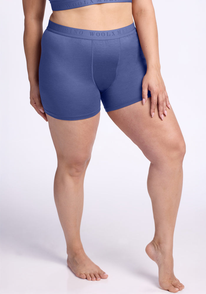 A person wears the Woolx Elliott Boyshort in Cloudy Bay with a matching blue sports bra, standing barefoot against a light background and resting one hand on their thigh. 