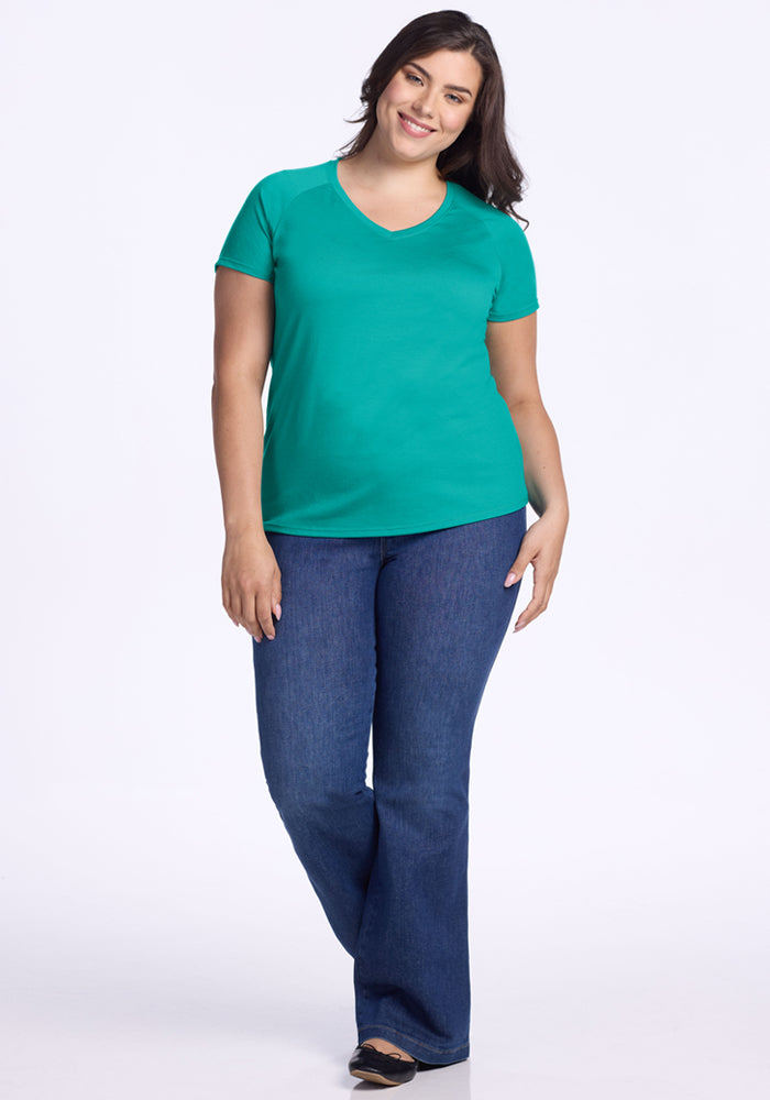 A woman with long dark hair smiles while wearing the Woolx Mia V Neck - Cool Jade, a bright green women's v-neck t-shirt made from moisture-wicking Merino wool, paired with blue jeans and black shoes against a white background.