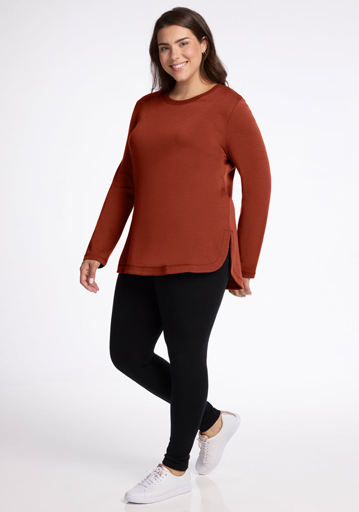 A woman wearing the Woolx Blair Tunic in Maple Leaf, paired with black leggings and white sneakers, stands smiling against a plain white background.