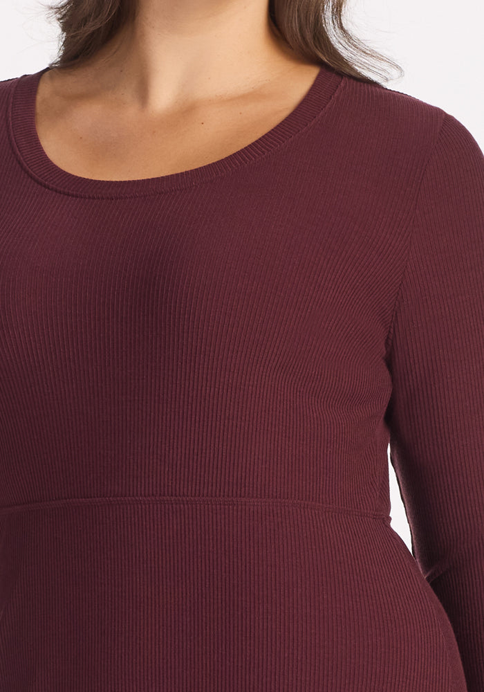A close-up of a person wearing the Woolx Caroline Ribbed Twirl Dress in Toasted Garnet, featuring a scoop neckline and versatile silhouette. Only the upper torso and part of the arm are visible against a white background.