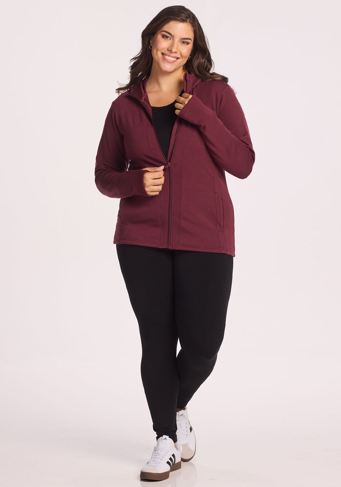 A woman with long brown hair smiles while wearing a Woolx Zoey Zip Up Hoodie in Toasted Garnet, a black top, black leggings, and white sneakers. She stands against a plain white background.