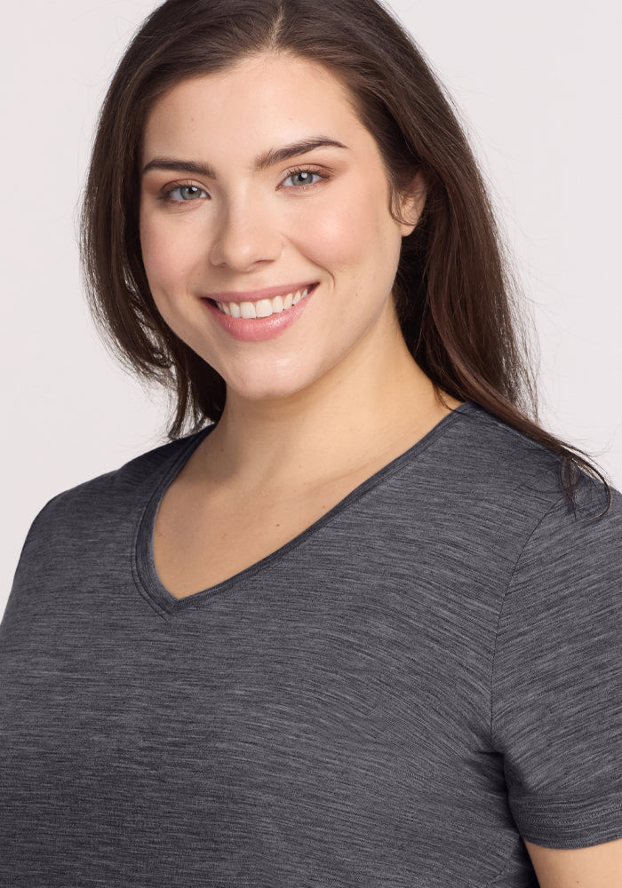 A woman with long brown hair smiles at the camera, wearing the Woolx Junie V Neck in Cinder Melange—a women's relaxed fit, short-sleeved Merino wool tee—against a light, plain background.